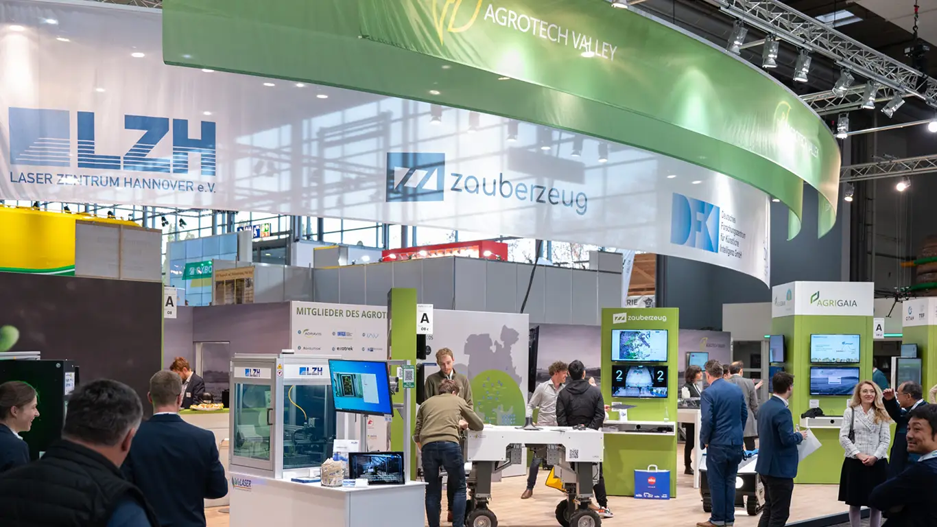 Blick auf den vollständigen Stand des Agrotech Valley auf der Agritechnica. Zahlreiche Besucher im Gespräch, Technik-Demonstrationen, Screens und Banner von Partnern wie LZH, DFKI, Zauberzeug und Agri-Gaia.